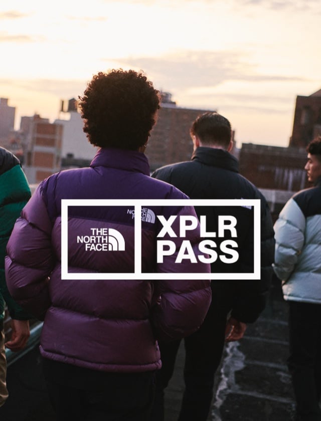 The XPLR Pass logo overlays an image of two people in Nuptse jackets watching a sunset over a city.
