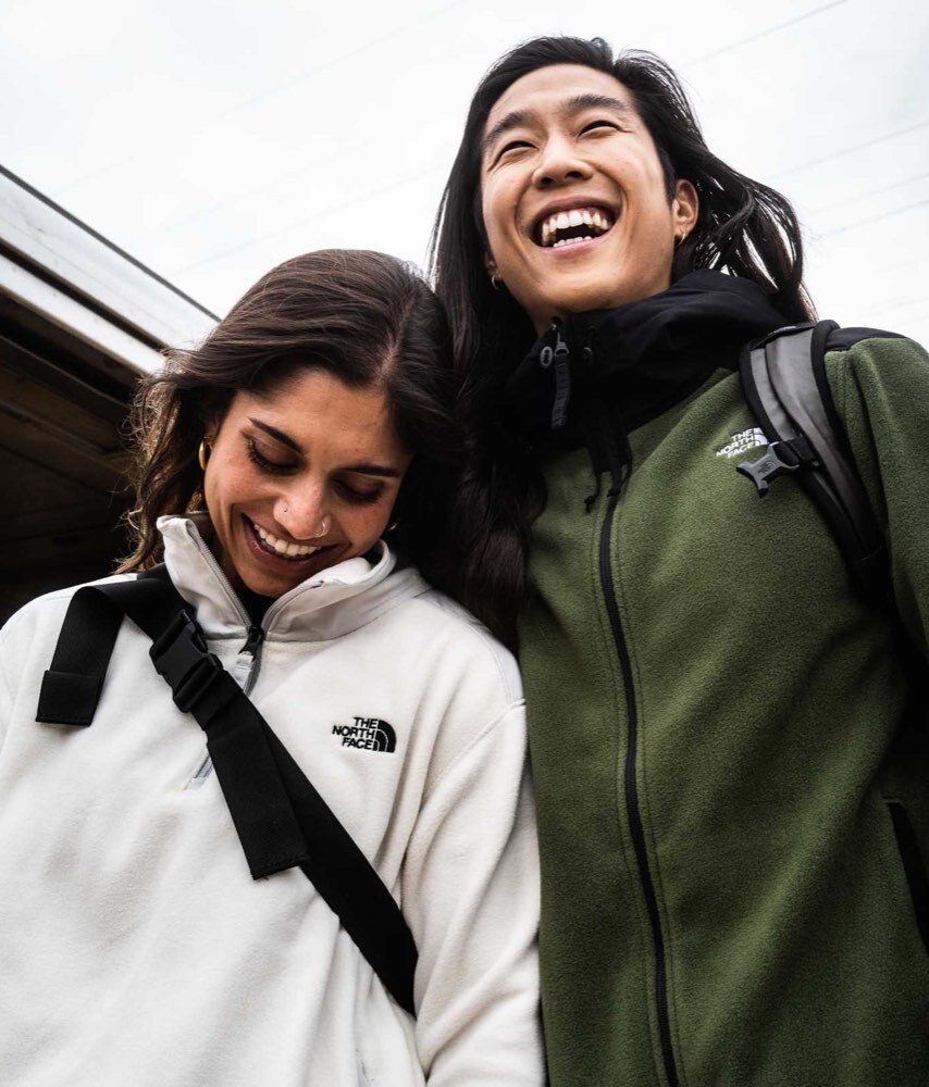 Two people wearing circular fleece from The North Face are smiling outside on a cloudy day.