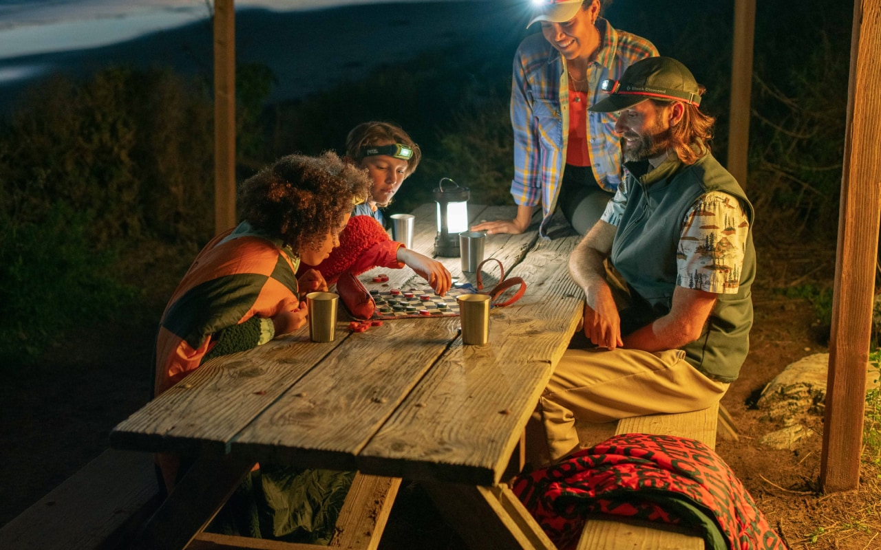 A family, wearing head lamps, plays checkers at an outdoor table as night falls.
