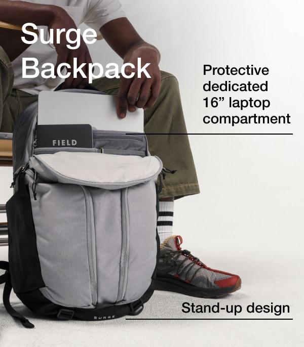 A person at a desk pulls a laptop from a gray Surge backpack.