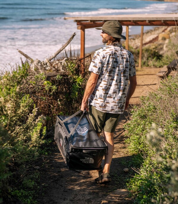 A camper carries a rugged bag out along a dirt path bordered by greenery.