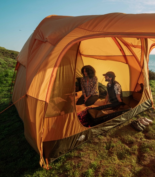 Two campers sit inside a spacious tent, pitched on a grassed area near the ocean.