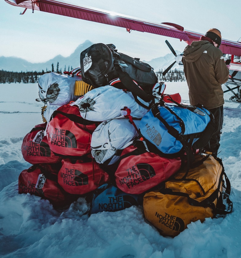 A stack of Base Camp Duffels in red and blue on a snowy landscape in front of a small plane.