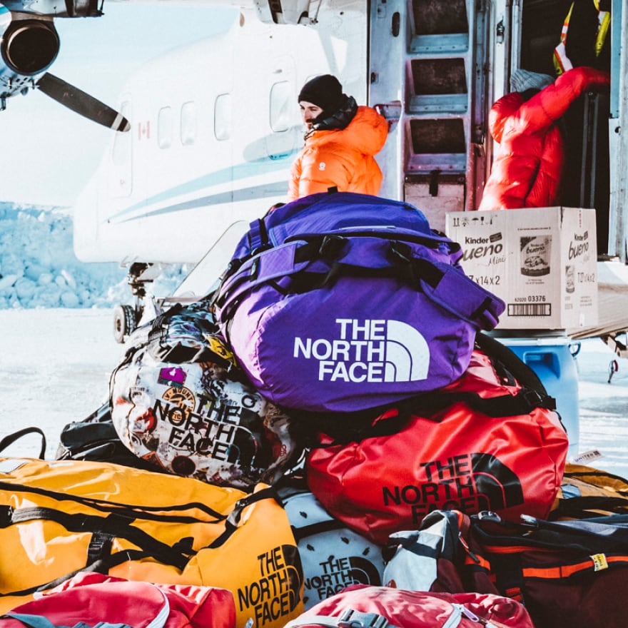 A stack of Base Camp Duffels in red and blue on a snowy landscape in front of a small plane.
