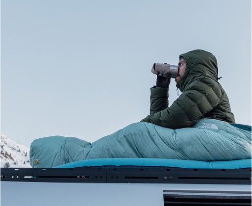 Person sitting in sleeping bag on top of van with mountains in the background.