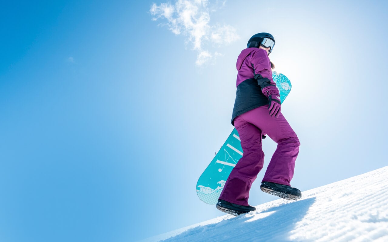 A woman wearing gear from the Freedom Collection carries her snowboard up the slopes on a clear day.