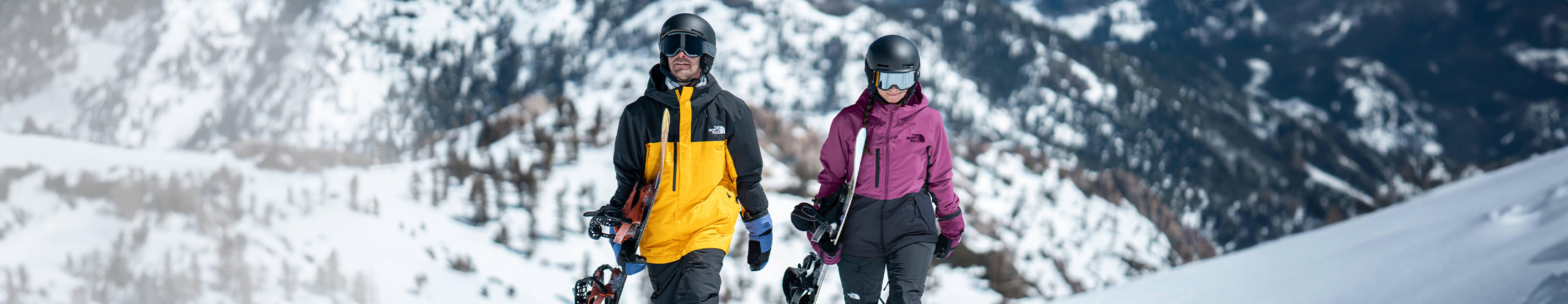 Two snowboarders wearing gear from The North Face carry their boards through a snowy mountain landscape.