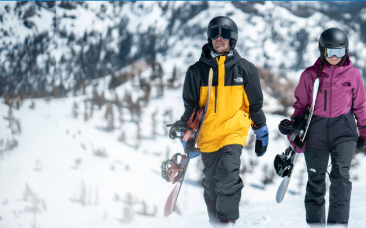 Deux planchistes vêtus d’équipement de The North Face transportent leurs planches dans un paysage de montagne enneigé.