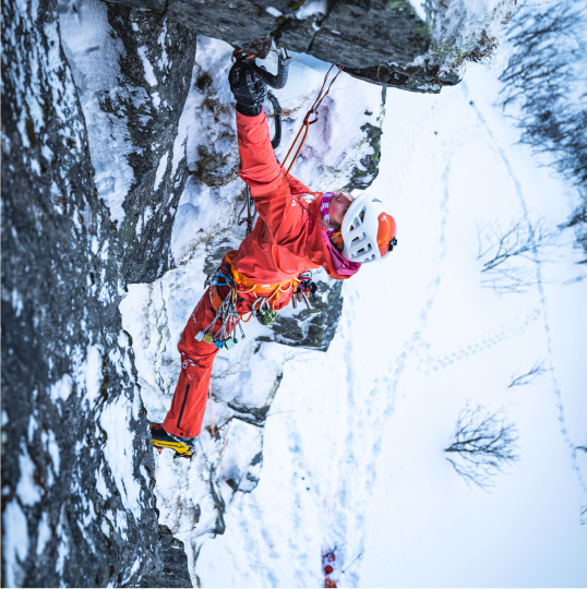 An athlete in a red expedition suit ice climbs in a wintry landscape.