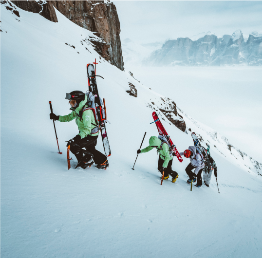 Three athletes bootpack up a snowy mountain with skis strapped to their backs.