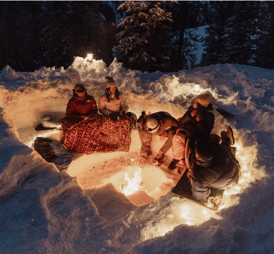 A group of friends huddles around a dug out campfire in the snow.