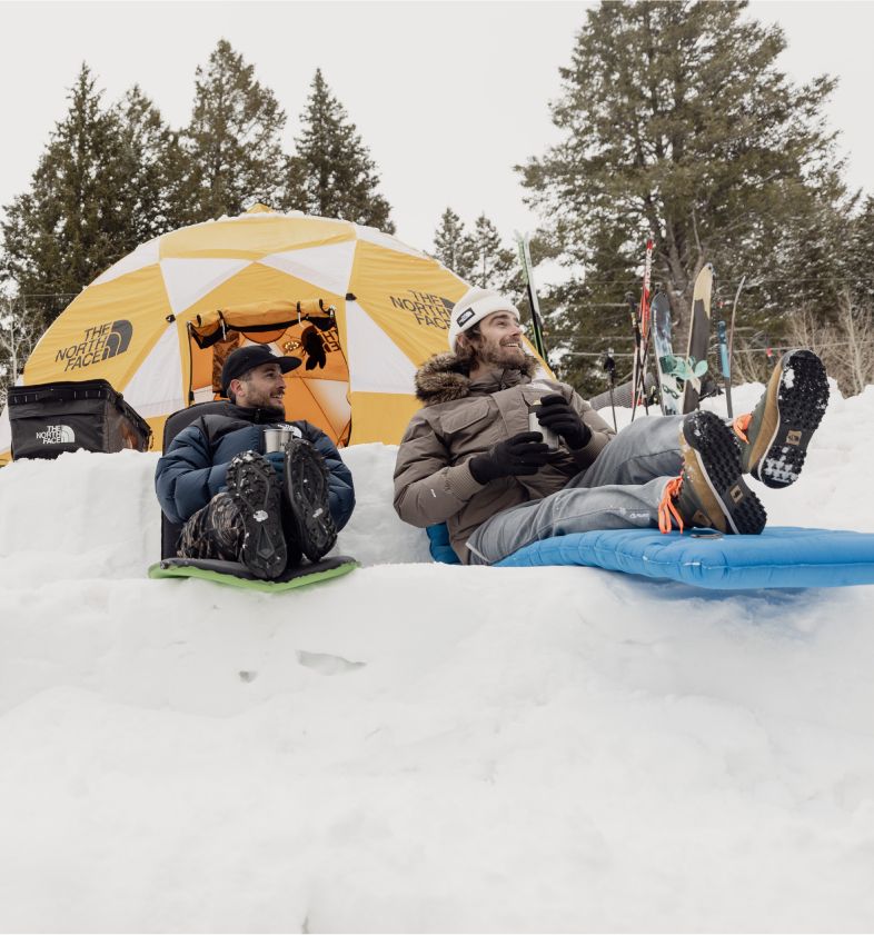 Two friends have set up camp in the snow. They are relaxing on their sleeping pads and holding their coffee outside their yellow tent. Their skis are set up in the distance.  