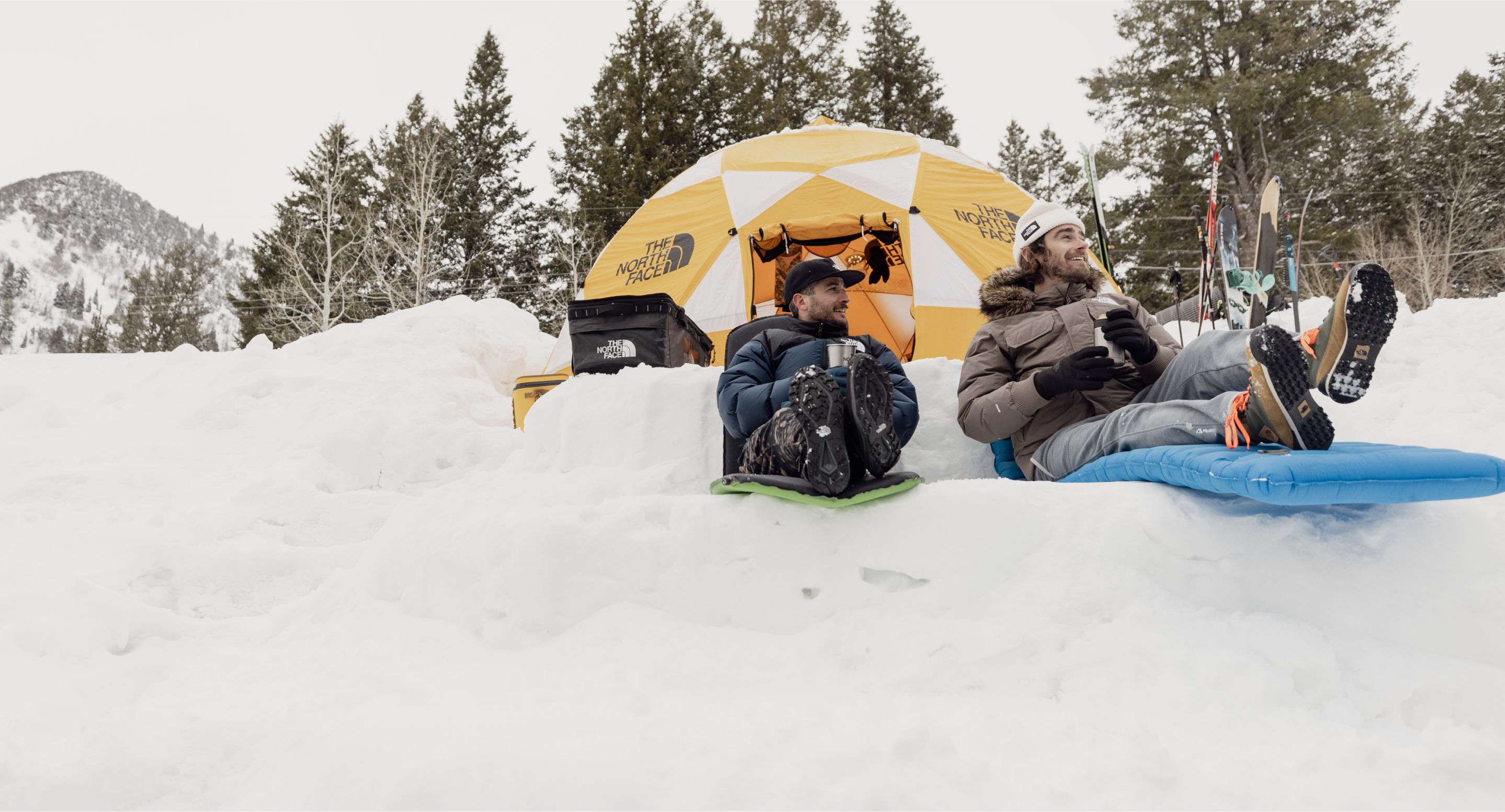 Two friends have set up camp in the snow. They are relaxing on their sleeping pads and holding their coffee outside their yellow tent. Their skis are set up in the distance.  