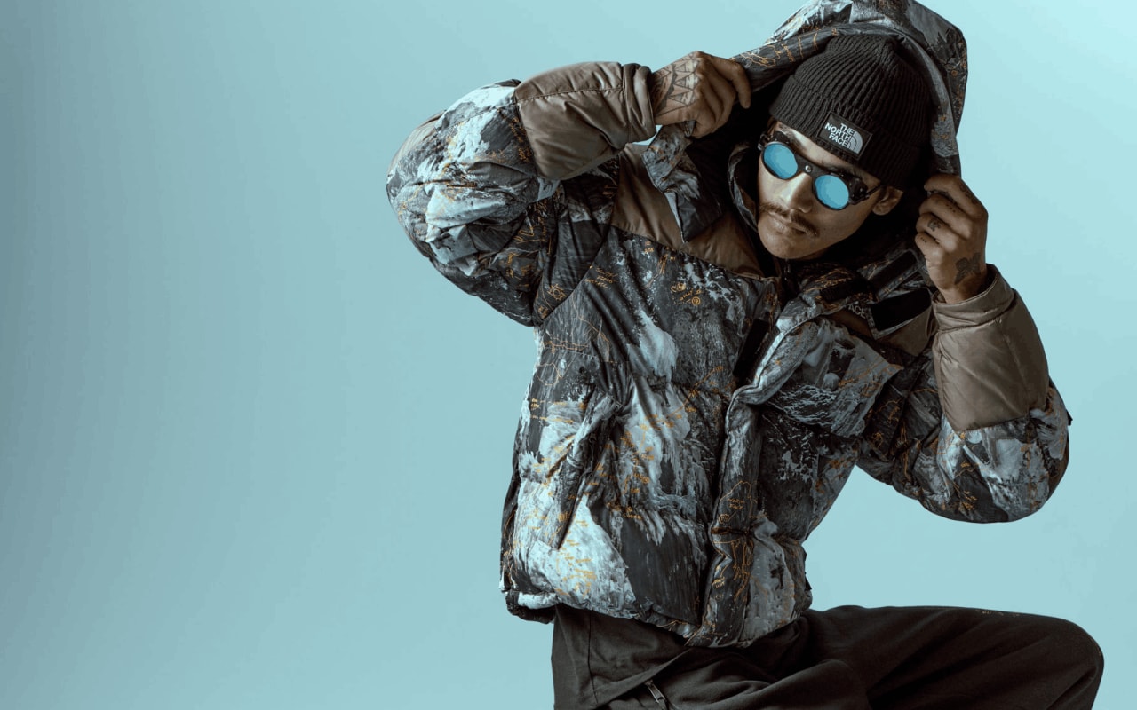 A person wearing the HMLYN Baltoro Jacket from Conrad Anker Notes Collection adjusts the hood in front of a blue background.
