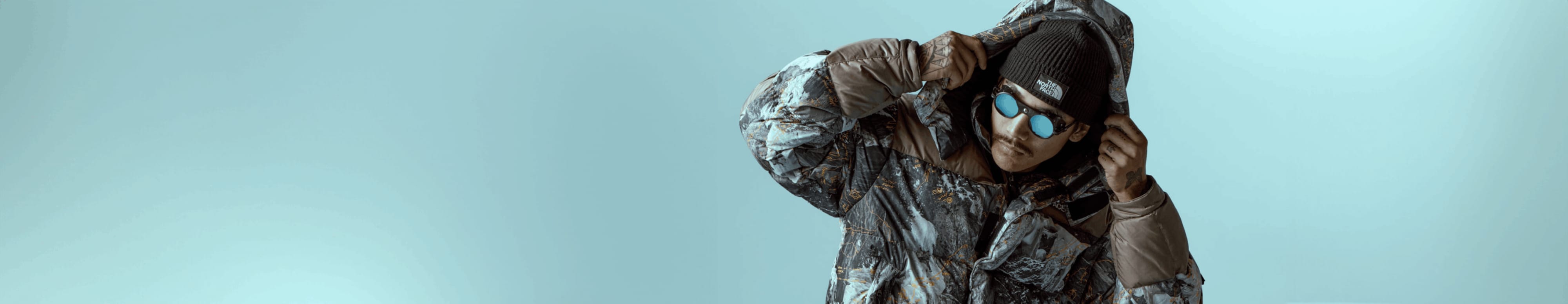 A person wearing the HMLYN Baltoro Jacket from Conrad Anker Notes Collection adjusts the hood in front of a blue background.
