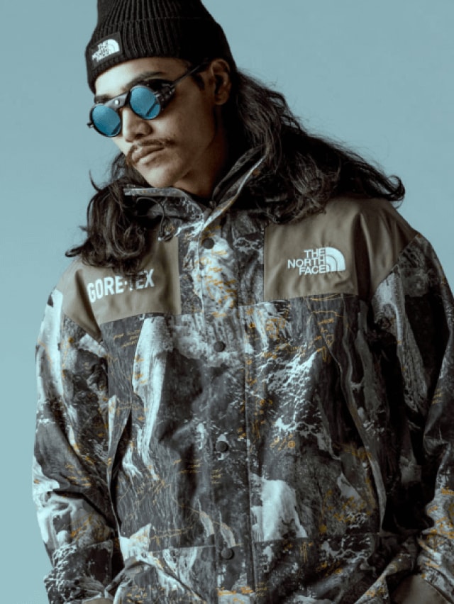 A person wearing the HMLYN Baltoro Jacket from Conrad Anker’s Notes Collection adjusts the hood in front of a blue background. 