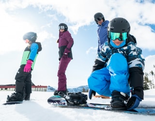 Snowboarder adjusting his bindings while his family waits to hit the slopes.