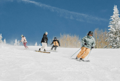 A group of four friends are doing a lap down the mountain. Two are snowboarding and two are skiing. They are wearing Freedom Insulated Bibs and Jackets by The North Face