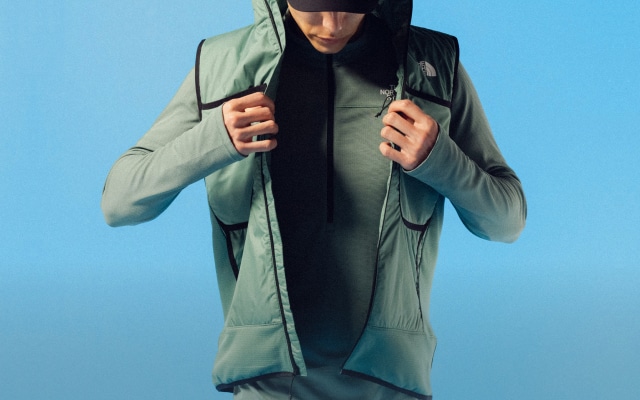 A man in cold weather running gear from The North Face puts on a jacket in a studio shot on a blue background. 