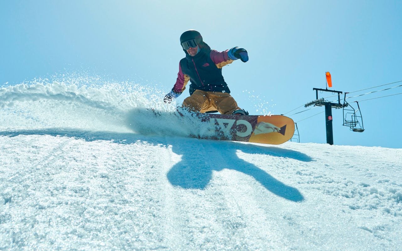 A person is kicking up snow on their snowboard wearing Men’s Freedom Insulated gear by The North Face. Text reads: “Conditions change. Weatherproof snow gear adapts.” 