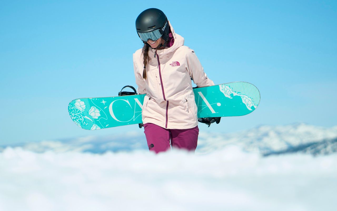A woman carrying a snowboard and wearing gear from The North Face hikes in the snow against a blue bird day.