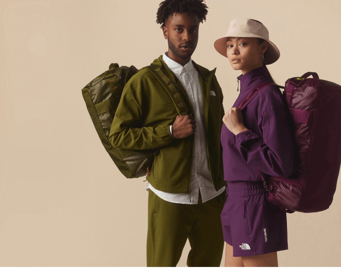 Studio shot of a man in all green Tekware from The North Face and a woman in all purple with Base Camp Voyagers on their backs. 