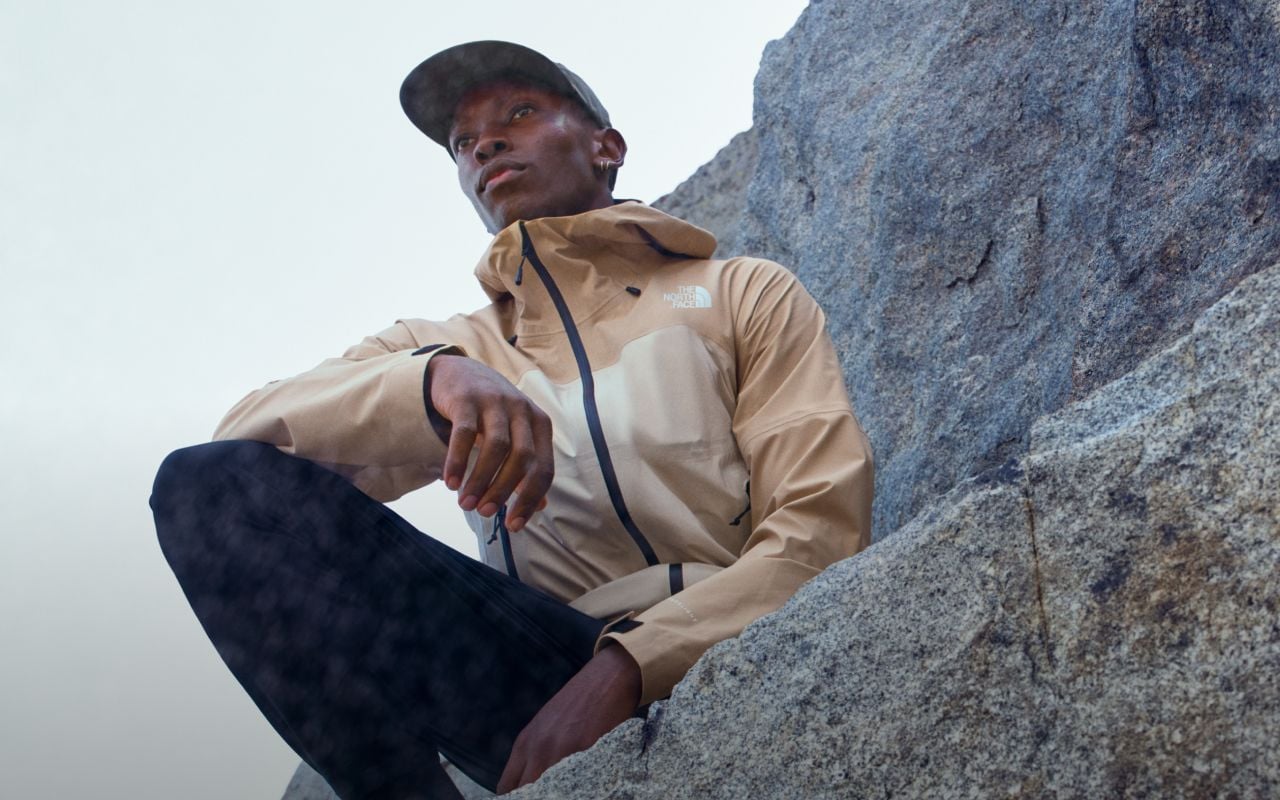 A person is sitting on a large rock outside looking into the distance. They are wearing a rain jacket from The North Face. Text reads: “Engineered for a life outside.” 