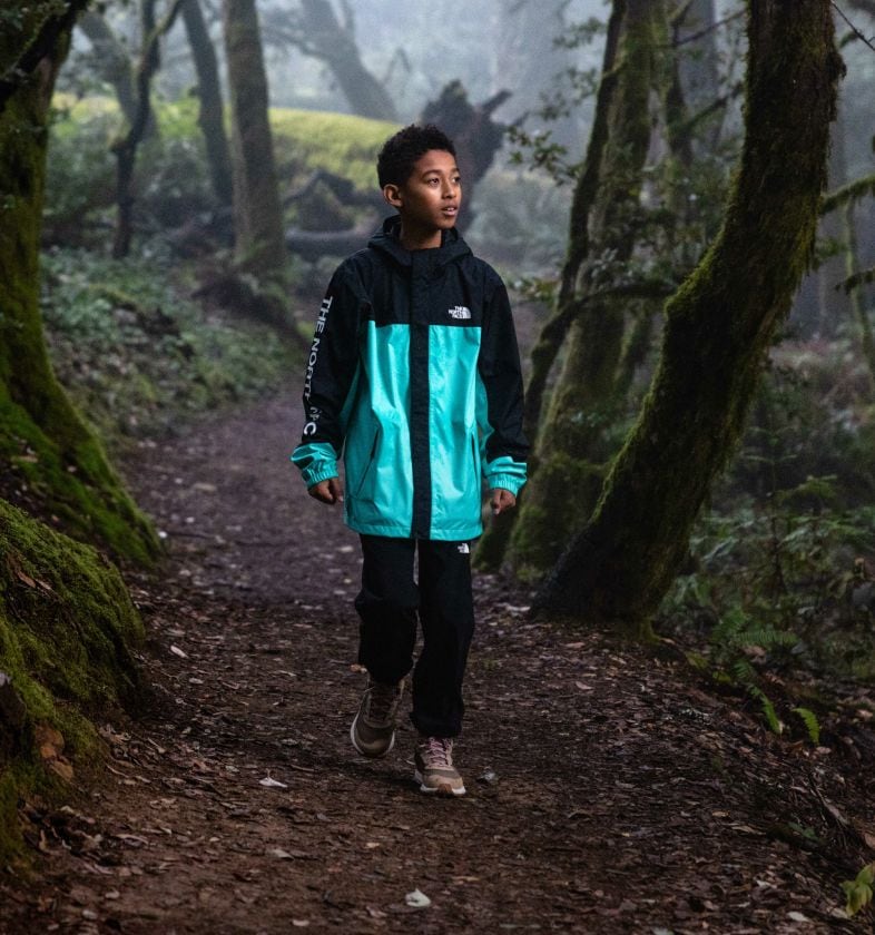 A child is walking in the woods wearing a black and teal-colored rain jacket from The North Face. Text reads: “Expand their horizons. Roam far and wide with your youngest explorers in these weather-ready favorites.” 