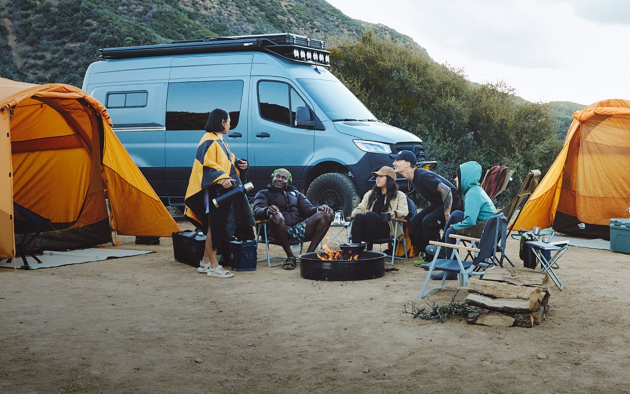 A group of friends are car camping. They’re bundled up sitting around the fire in front their large tents from The North Face. Text reads: “Not home. But there’s no place like this, either.” 