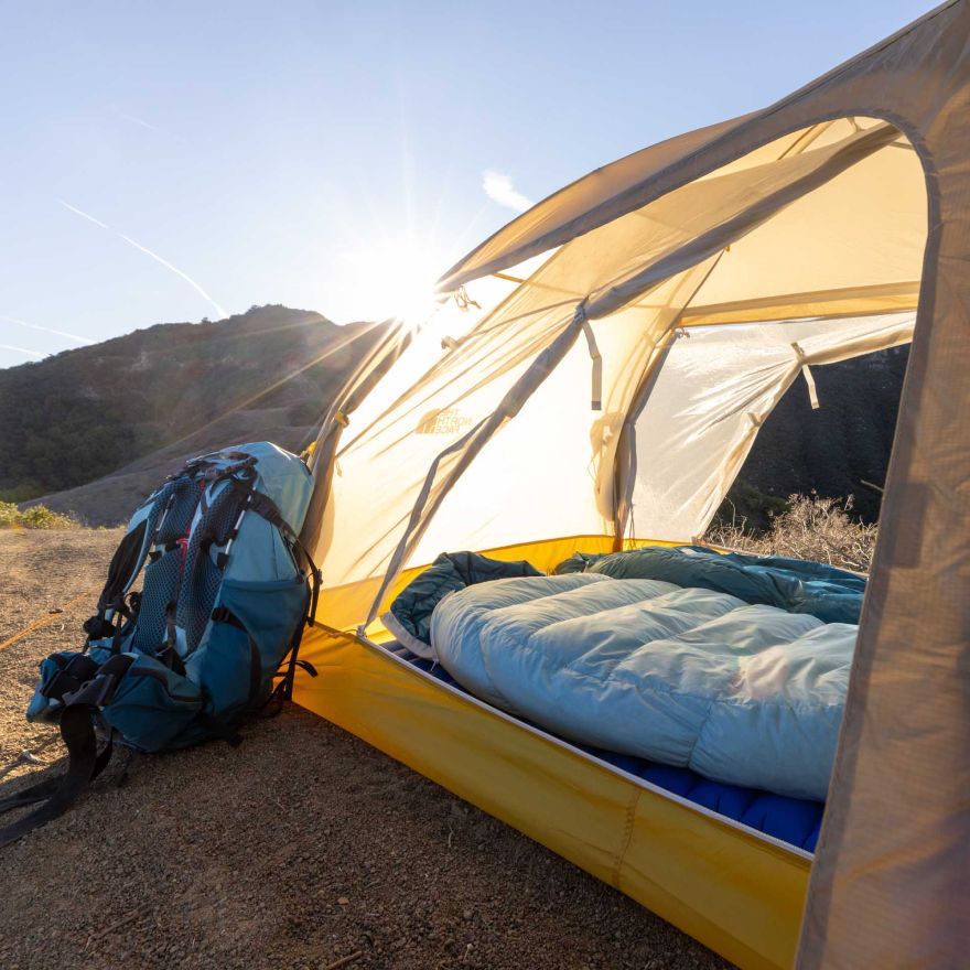 The sun is rising over the mountains. A backpack is leaning against a tent and sleeping bag, all from The North Face. 