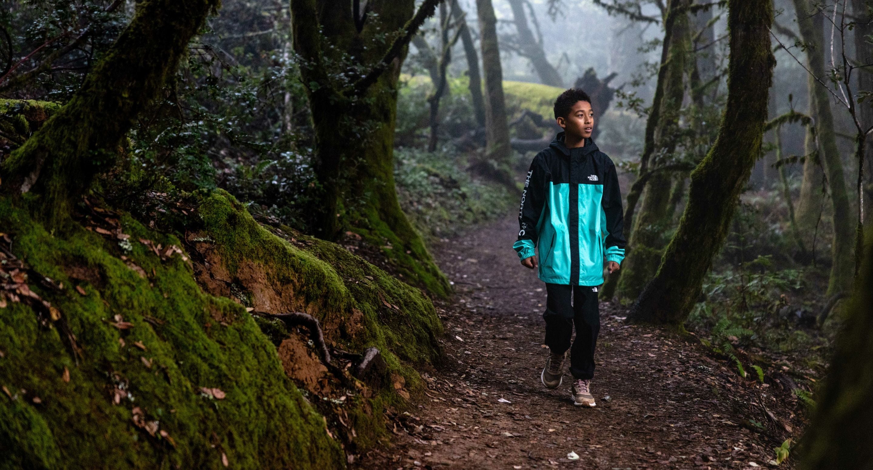 A child is walking in the woods wearing a black and teal-colored rain jacket from The North Face. Text reads: “Expand their horizons. Roam far and wide with your youngest explorers in these weather-ready favorites.” 
