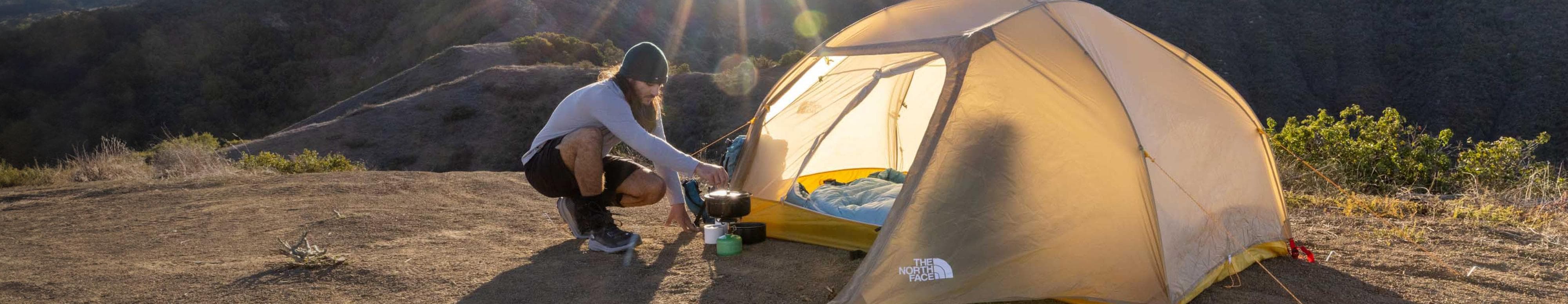 Une personne cuisine sur son réchaud de camping à l’extérieur de sa tente The North Face. La mention suivante y est inscrite : « Get back to the basics ». 