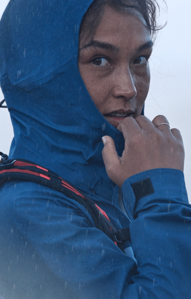 A hiker adjusts their hood while wearing a blue Frontier FUTURELIGHT™ Jacket in rainy conditions.