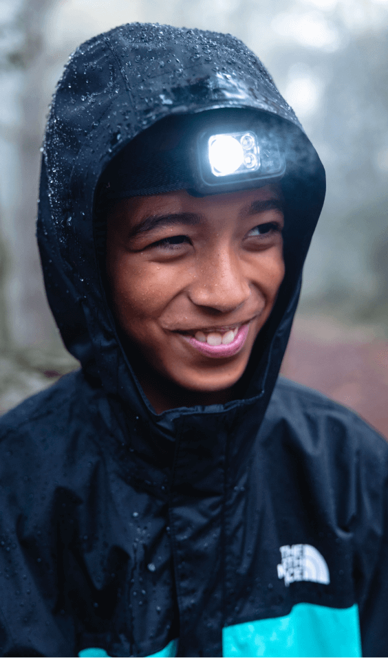 A kid wearing a waterproof jacket smiles while walking through the forest with a headlamp. 