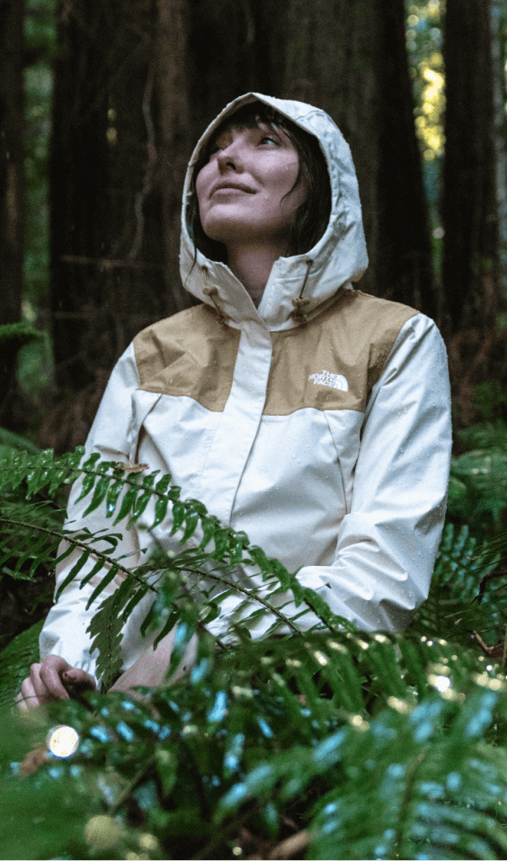 A woman sits in a rainy forest while wearing a waterproof jacket with the hood up.  