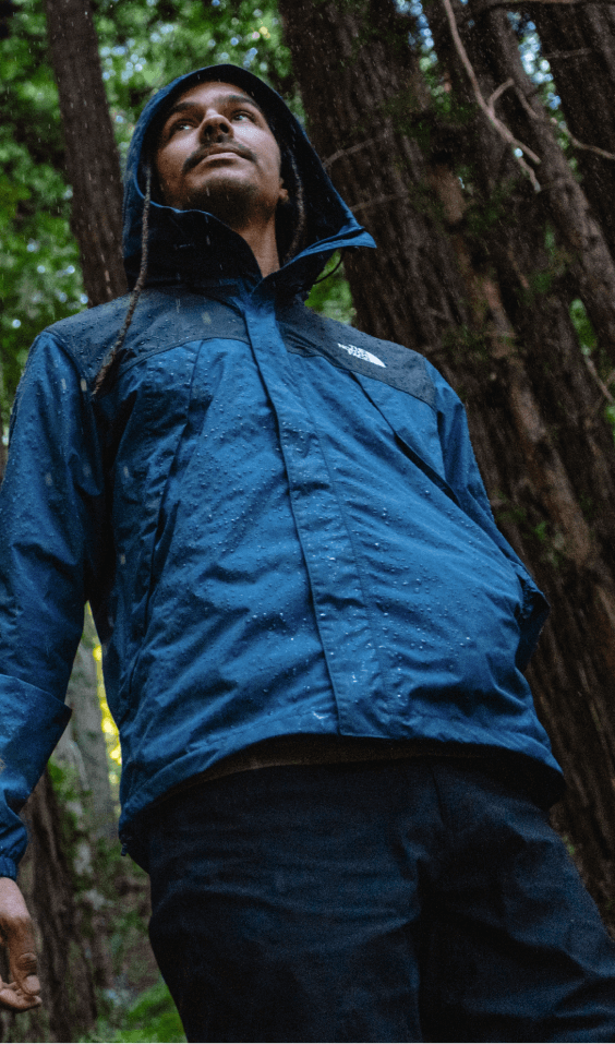 A man walks through a rainy forest as droplets shed off his waterproof jacket. 