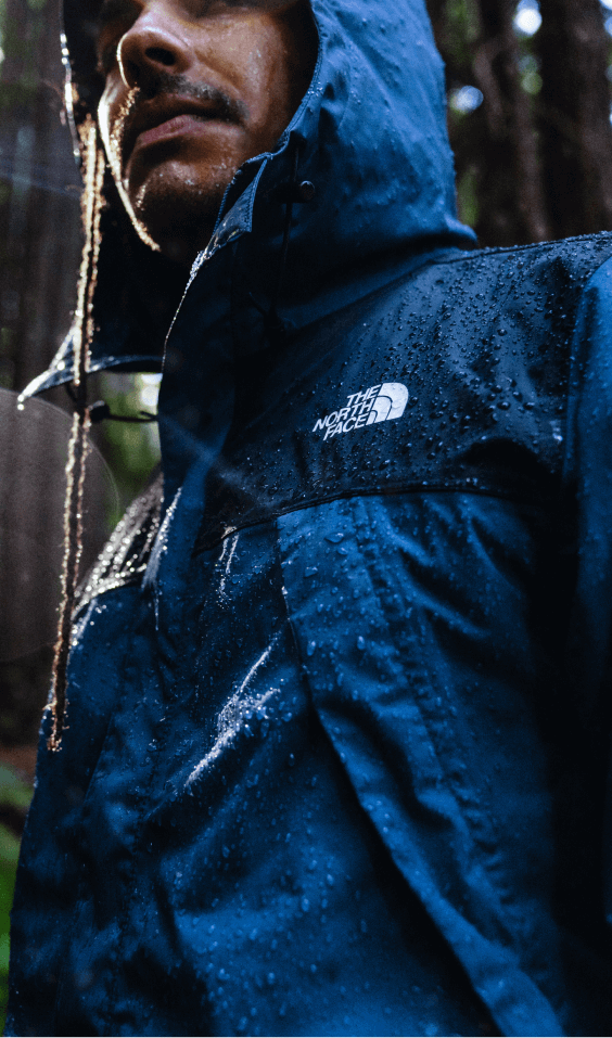 Un homme marche dans une forêt pluvieuse; des gouttelettes perlent sur sa veste imperméable. 