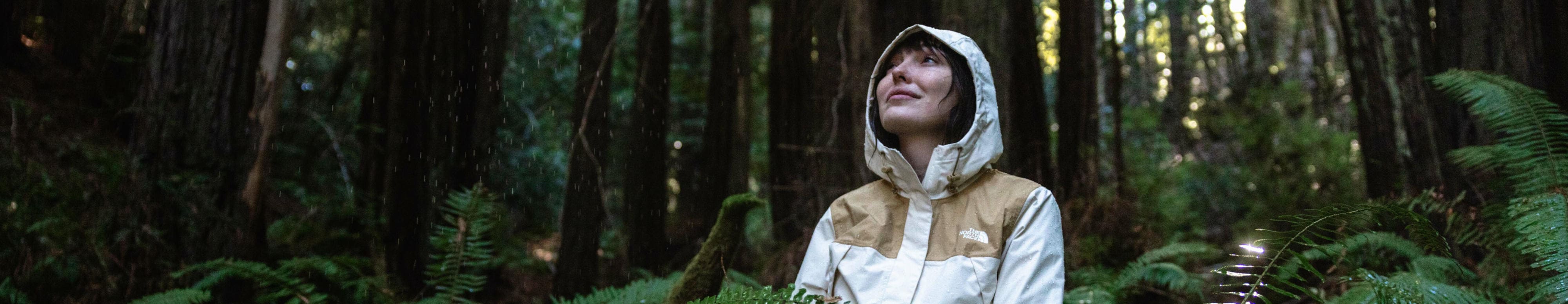 A woman sits in a rainy forest while wearing a waterproof jacket with the hood up. 