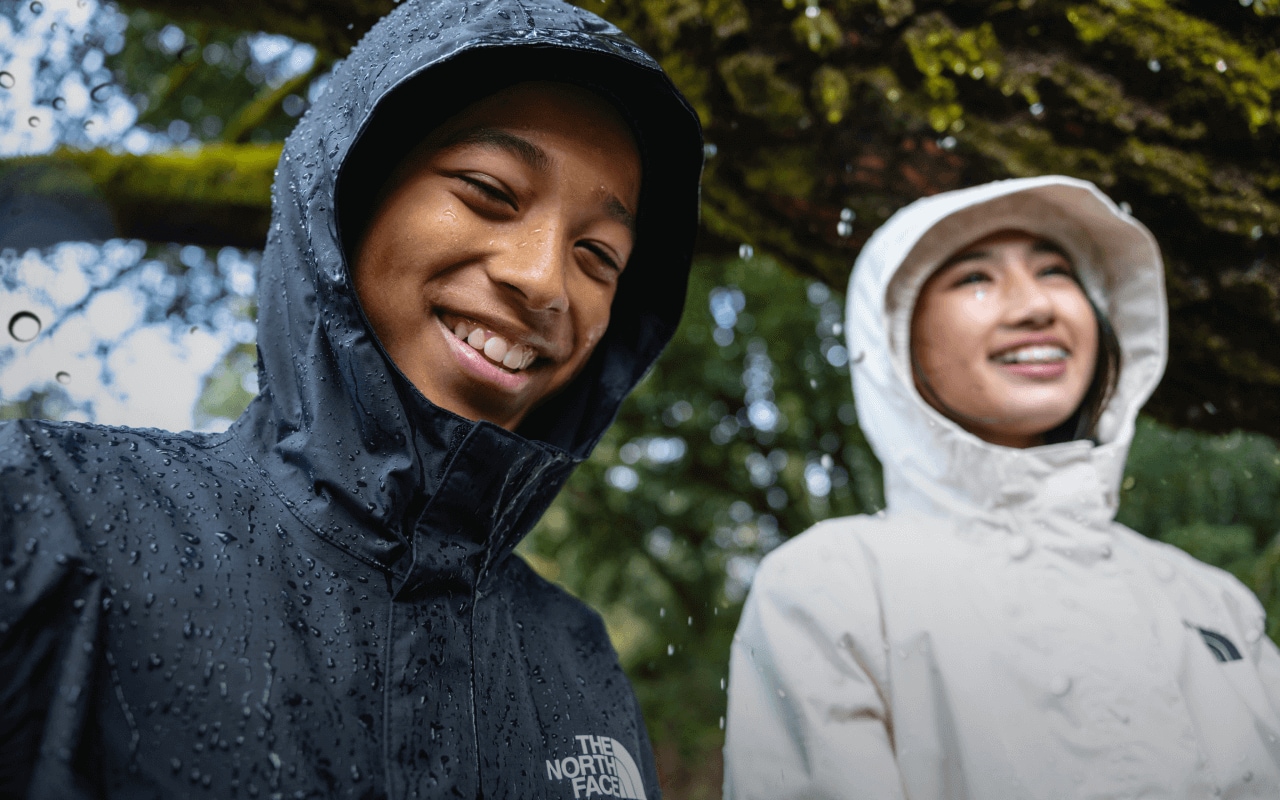 Two kids wearing waterproof jackets sit on a large, moss-covered tree branch. 