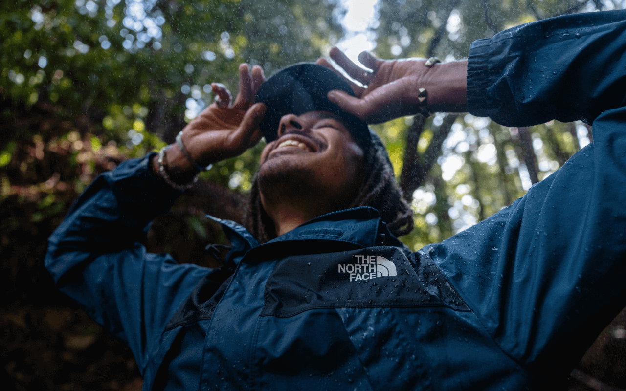 A man wearing a waterproof jacket stands with his hands in his pocket in the middle of a rainy forest.