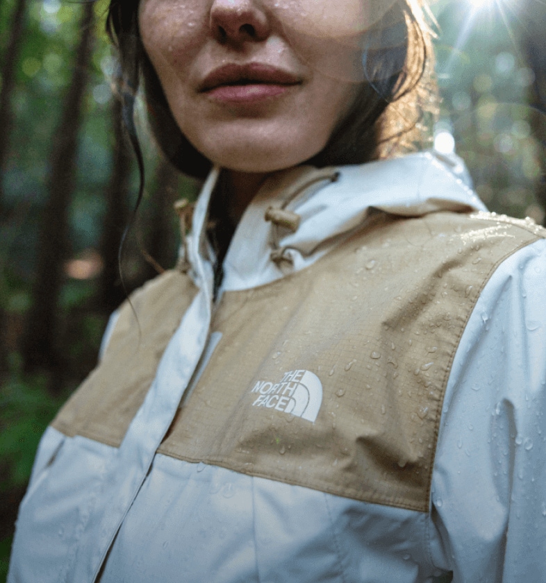 Une femme marche dans une forêt pluvieuse; des gouttelettes perlent sur sa veste imperméable.