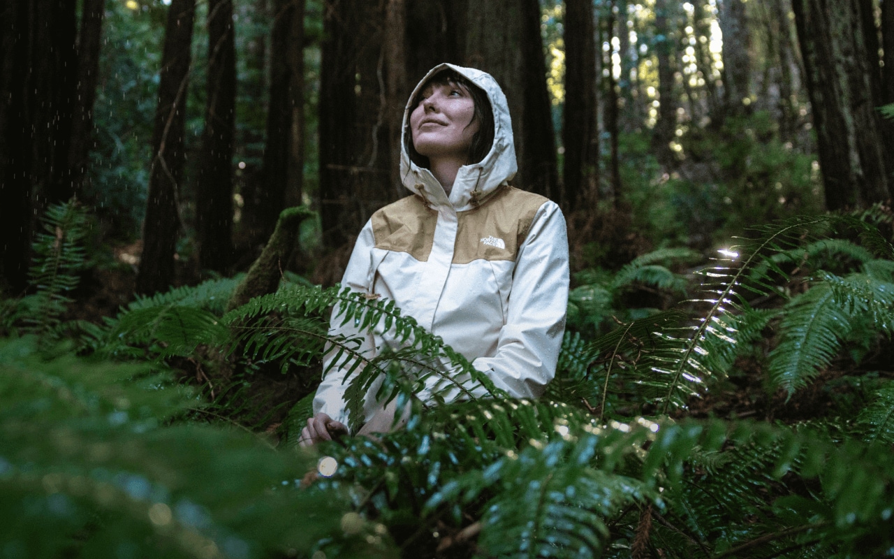 Une femme portant une veste imperméable avec la capuche relevée est assise dans une forêt pluvieuse.