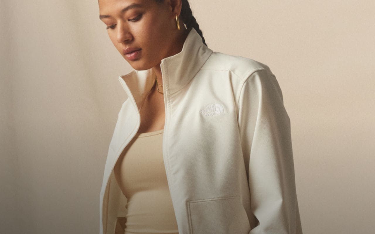 A woman is looking down wearing an unzipped Willow Stretch jacket in White Dune and a light brown beanie. Text reads: “Protective Willow Stretch jackets go wherever the wind takes you.” 