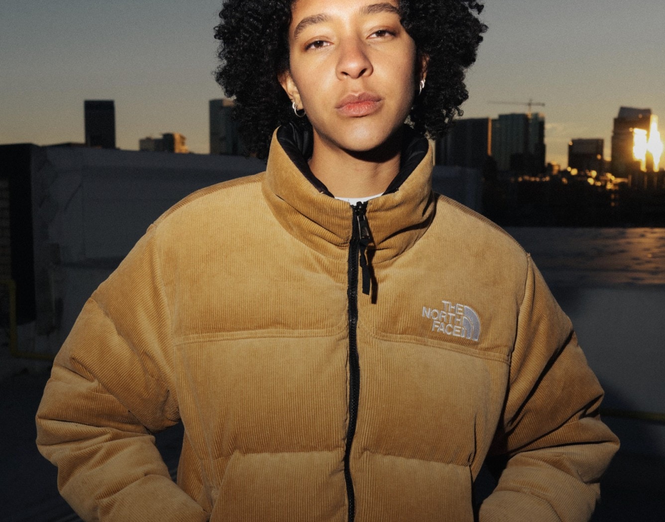 A person wears a Nuptse Jacket in beige against a city skyline.