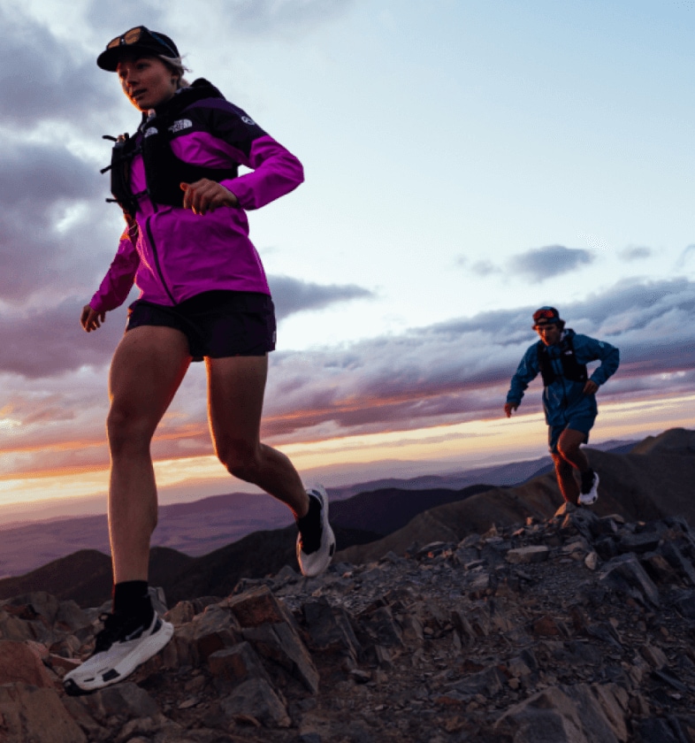 Deux athlètes The North Face courant sur des sentiers rocailleux et techniques au lever du soleil.