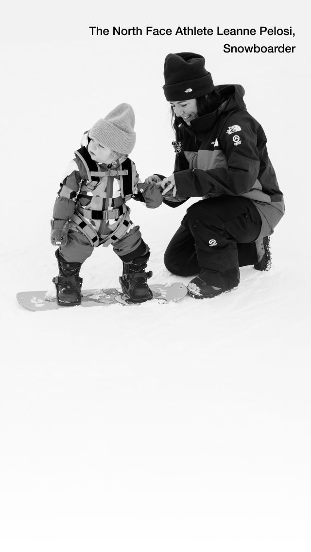 Black and white image of The North Face Athlete Leanne Pelosi kneeling in the snow helping her toddler with their snowsuit.