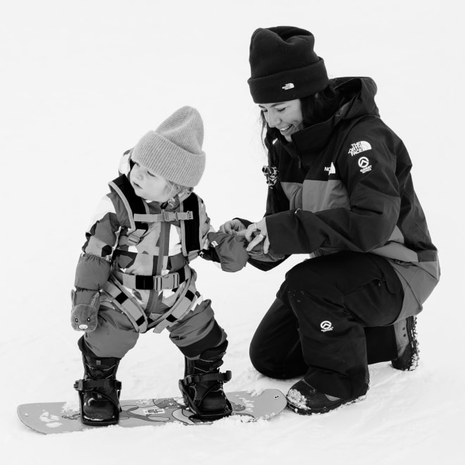 Black and white image of The North Face Athlete Leanne Pelosi kneeling in the snow helping her toddler with their snowsuit.