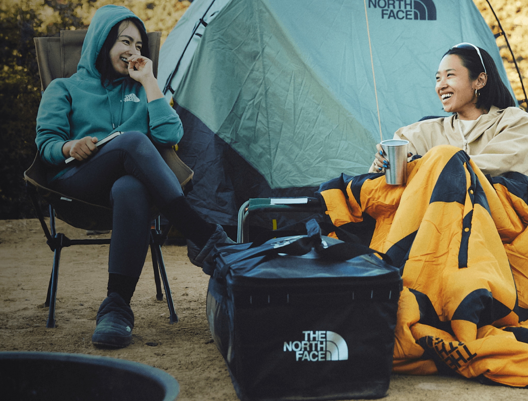 Two people sit on camp chairs at camp, laughing under The North Face blankets. 
