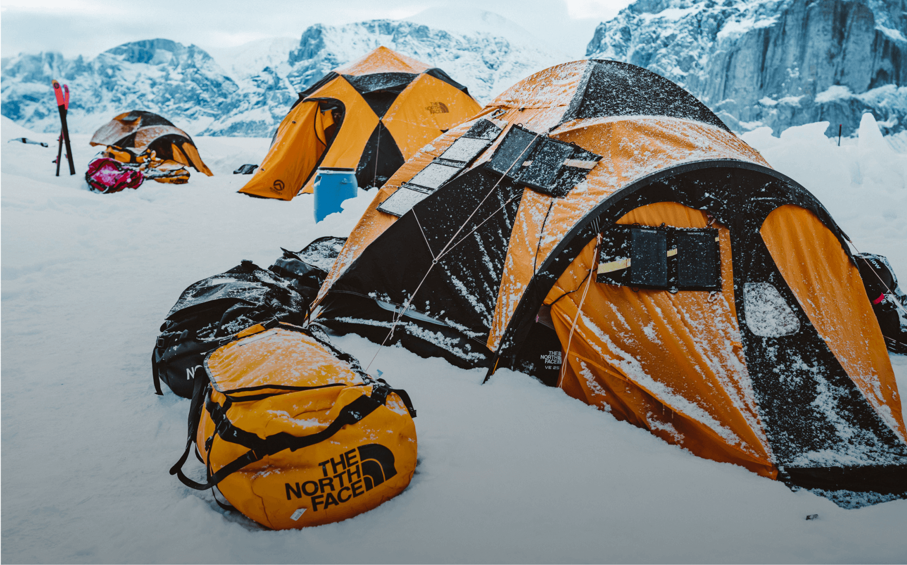 A base camp of tents at the foot of jagged, snowy peaks. 