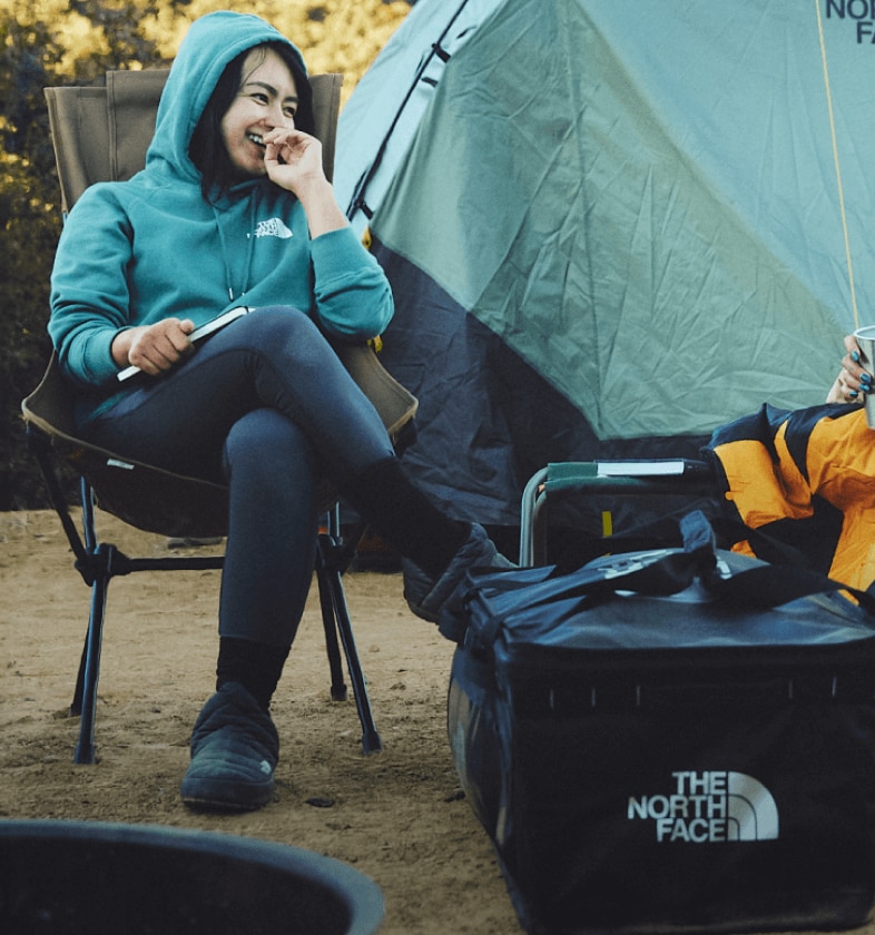 Two people sit on camp chairs at camp, laughing under The North Face blankets. 